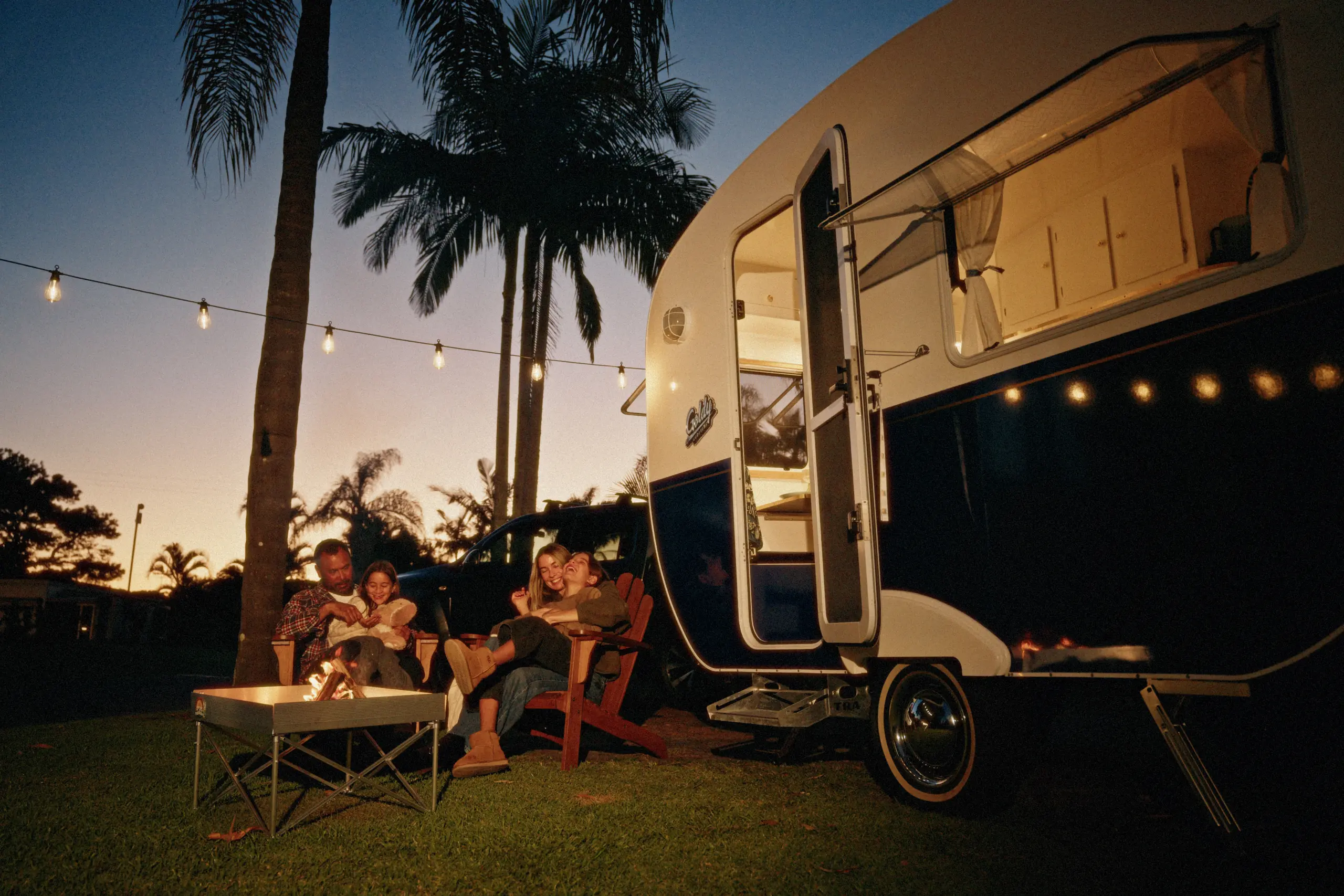 Family sits around a campfire in front of a retro camper at dusk, string lights overhead, palm trees, outdoor cozy evening vibe.