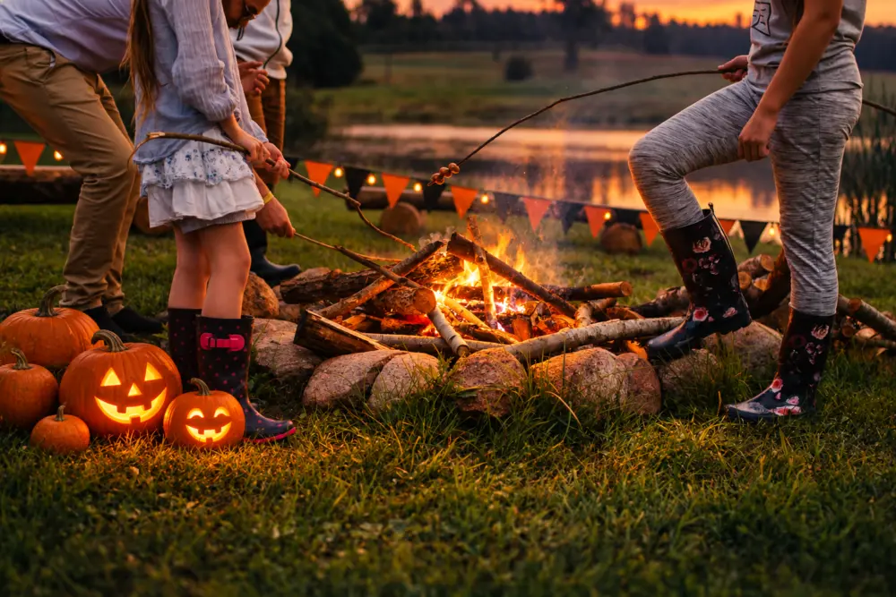 Children and adults tend a bonfire at dusk, carving sticks, glowing jack-o’-lanterns on the grass, pumpkins nearby, lake in the background.