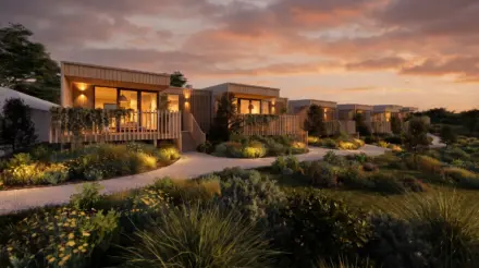 Row of modern wooden cabins with warm lights, elevated decks, lush desert-inspired landscaping, winding gravel paths, and a sunset sky.