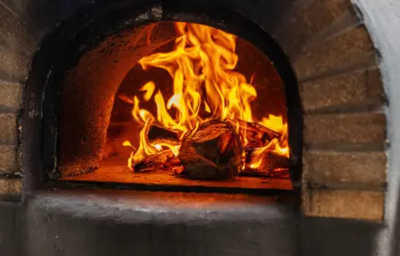 A close‑up of a glowing wood fire inside a brick oven, with orange flames curling around logs and a dark, sooty interior.