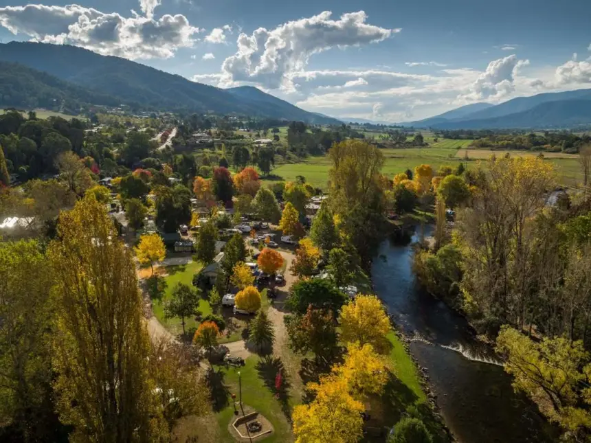 Aerial view of a valley town with colorful autumn trees, a winding river, and a cluster of homes and campers along a park-like riverbank, mountains in the distance.
