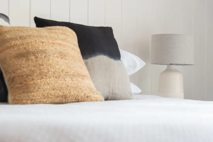 Soft beige, black, and cream cushions resting on a light bedspread with a neutral table lamp against a white paneled wall.