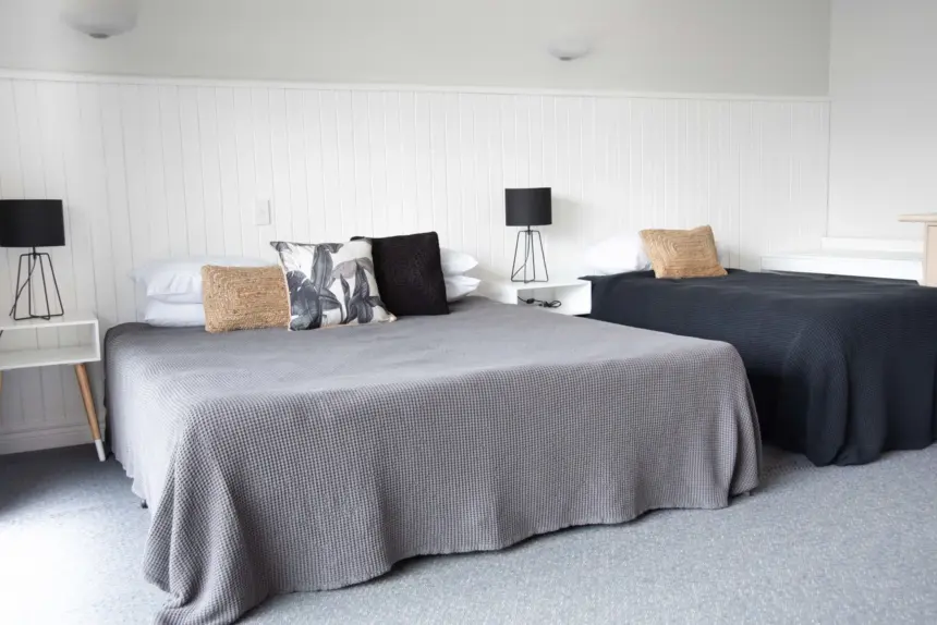 Two neatly made beds with grey and navy covers, white panelled wall, black lamps on white nightstands, and neutral cushions in a bright, minimalist room.
