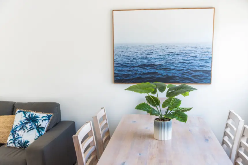 Cozy dining area with a wooden table and light chairs, a potted plant, a sea framed print on the wall, and a grey sofa with tropical cushions.