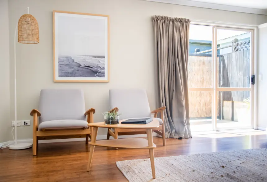 Cozy living room with two light grey cushioned armchairs, wooden frames, a round coffee table, a framed beach photo, woven floor lamp, and sliding glass door with beige curtains.
