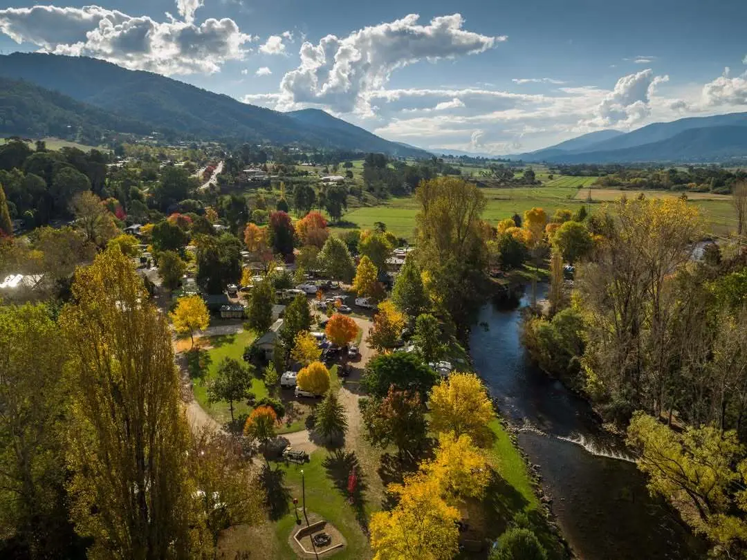 Aerial view of a valley town with colorful autumn trees, a winding river, and a cluster of homes and campers along a park-like riverbank, mountains in the distance.
