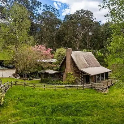 A rustic wooden cabin with a steep roof sits beside a fenced yard in a grassy, tree-filled clearing, with a pink flowering tree and blue sky above.