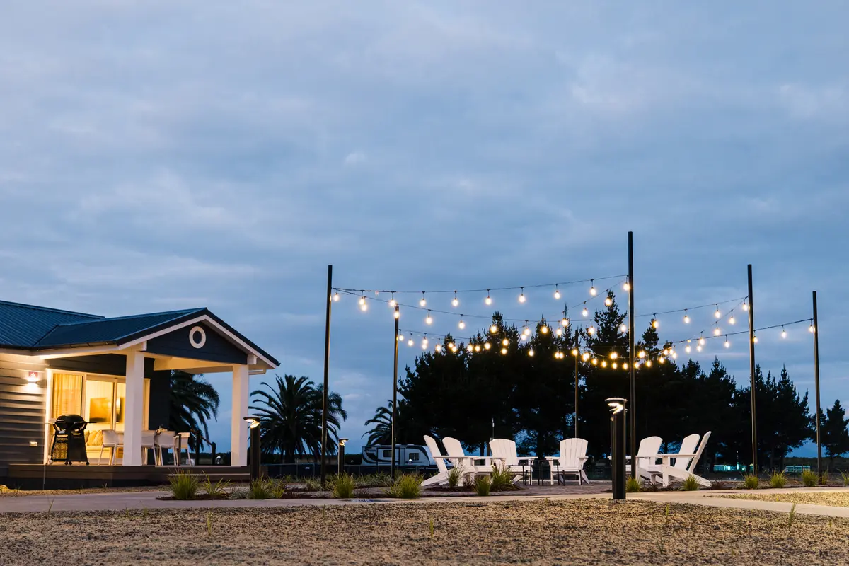 Outdoor evening scene at a modern house with string lights, white chairs, a small patio, palm trees in the background, and a sandy foreground.