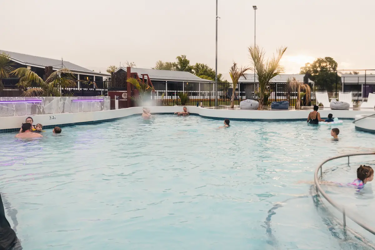 People enjoy a resort pool with gentle waves, palm trees, and lounge areas in the background under a soft sunset sky.
