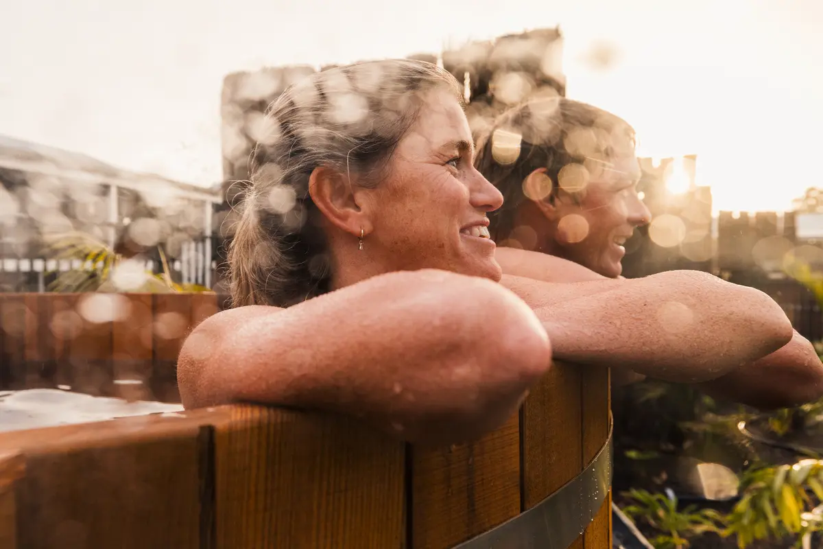 Two people lean on a wooden hot tub rim, smiling and chatting, warm sunset lighting and soft bokeh circles in the foreground.