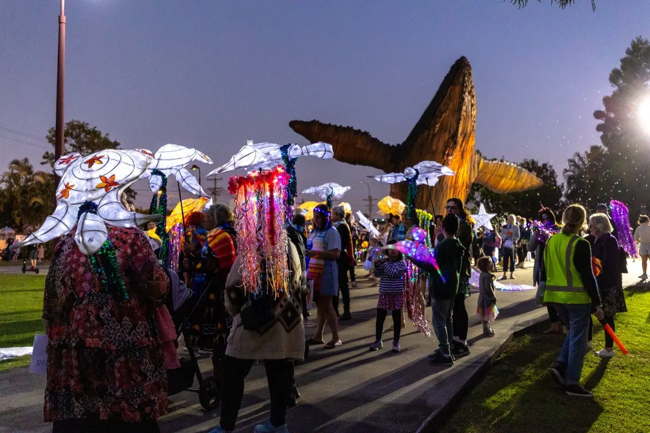 A lively evening parade with illuminated jellyfish lanterns, colorful costumes, and a giant glowing bird sculpture in a park, crowd chatting and enjoying the spectacle.