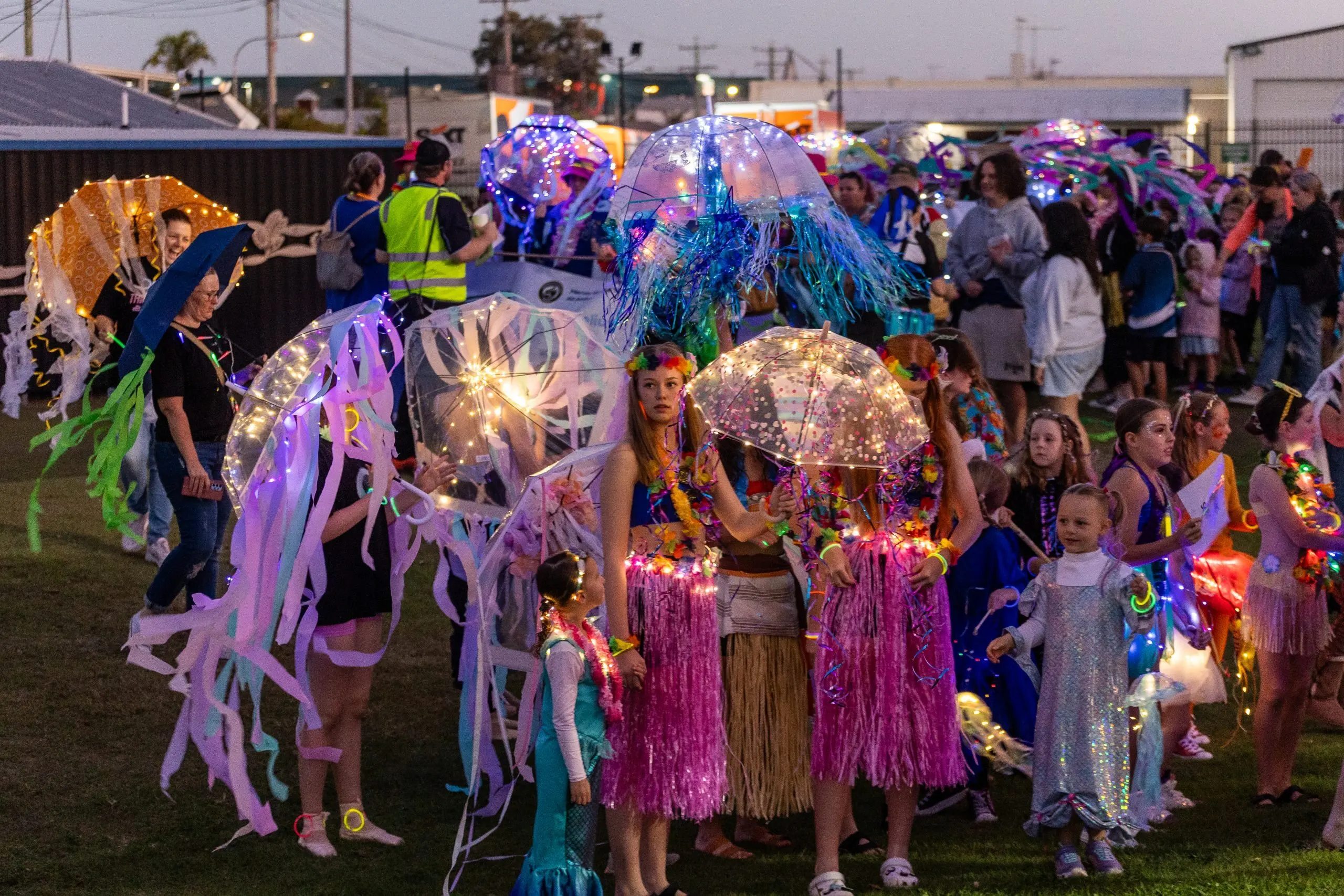 Colorful parade of kids in glowing, fringed costumes and illuminated umbrellas, twinkling lights, streamers, and festive outfits at dusk.
