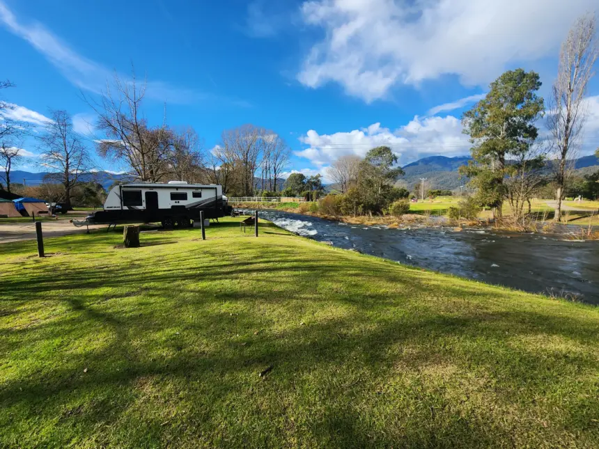 Scenic riverside campground with a white travel trailer parked on a grassy bank, a fast-flowing river, leafless trees, tents, and distant mountains under a bright blue sky.