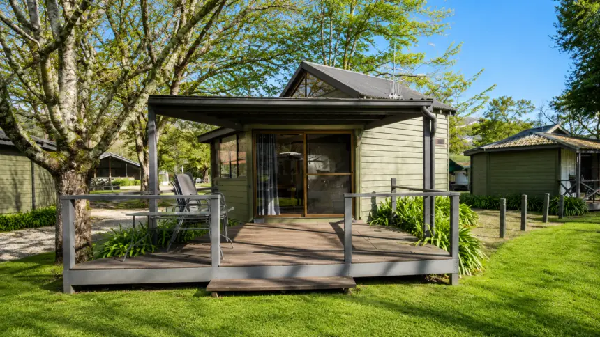 Small wooden cabin with a raised deck, sliding door, and chairs, surrounded by green lawn and trees in a sunny park-like setting.