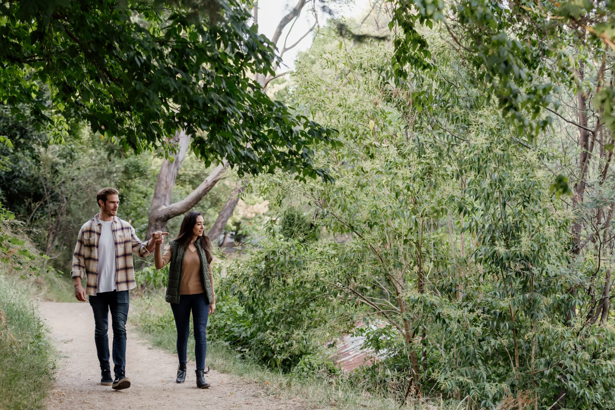 Two people walk along a leafy trail hand in hand under overhanging branches, surrounded by green trees and bushes in a serene park.