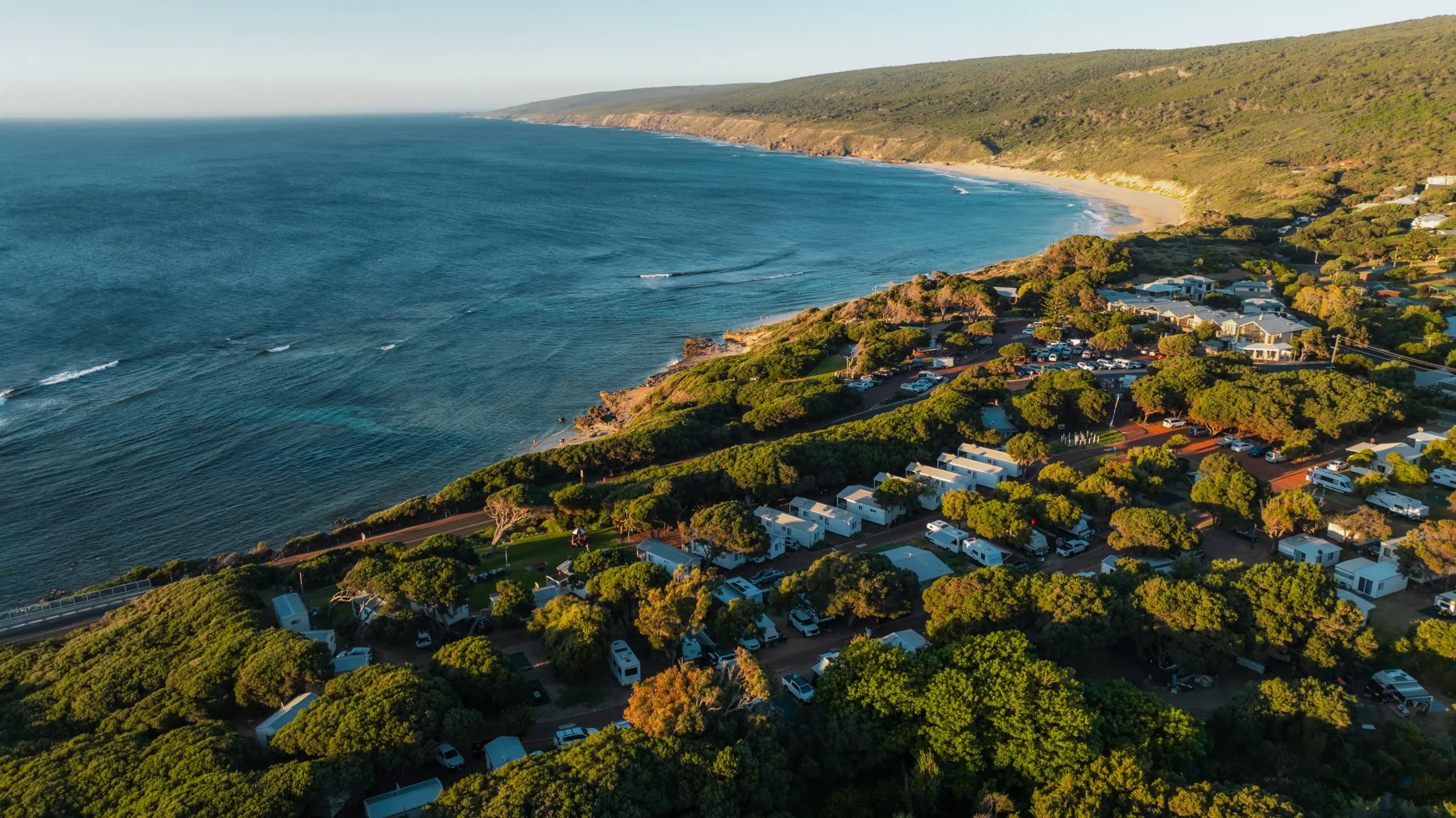 A coastal town with a blue ocean to the left, sandy beach and rocky shoreline, and a forested hillside dotted with houses, trees, and parked vehicles.