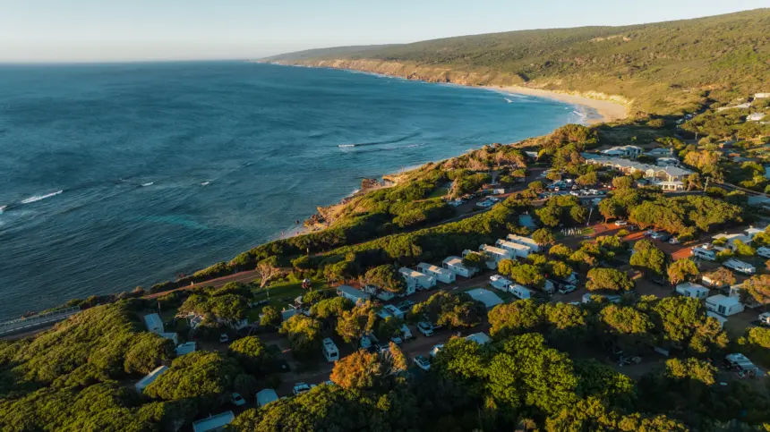 A coastal town with a blue ocean to the left, sandy beach and rocky shoreline, and a forested hillside dotted with houses, trees, and parked vehicles.
