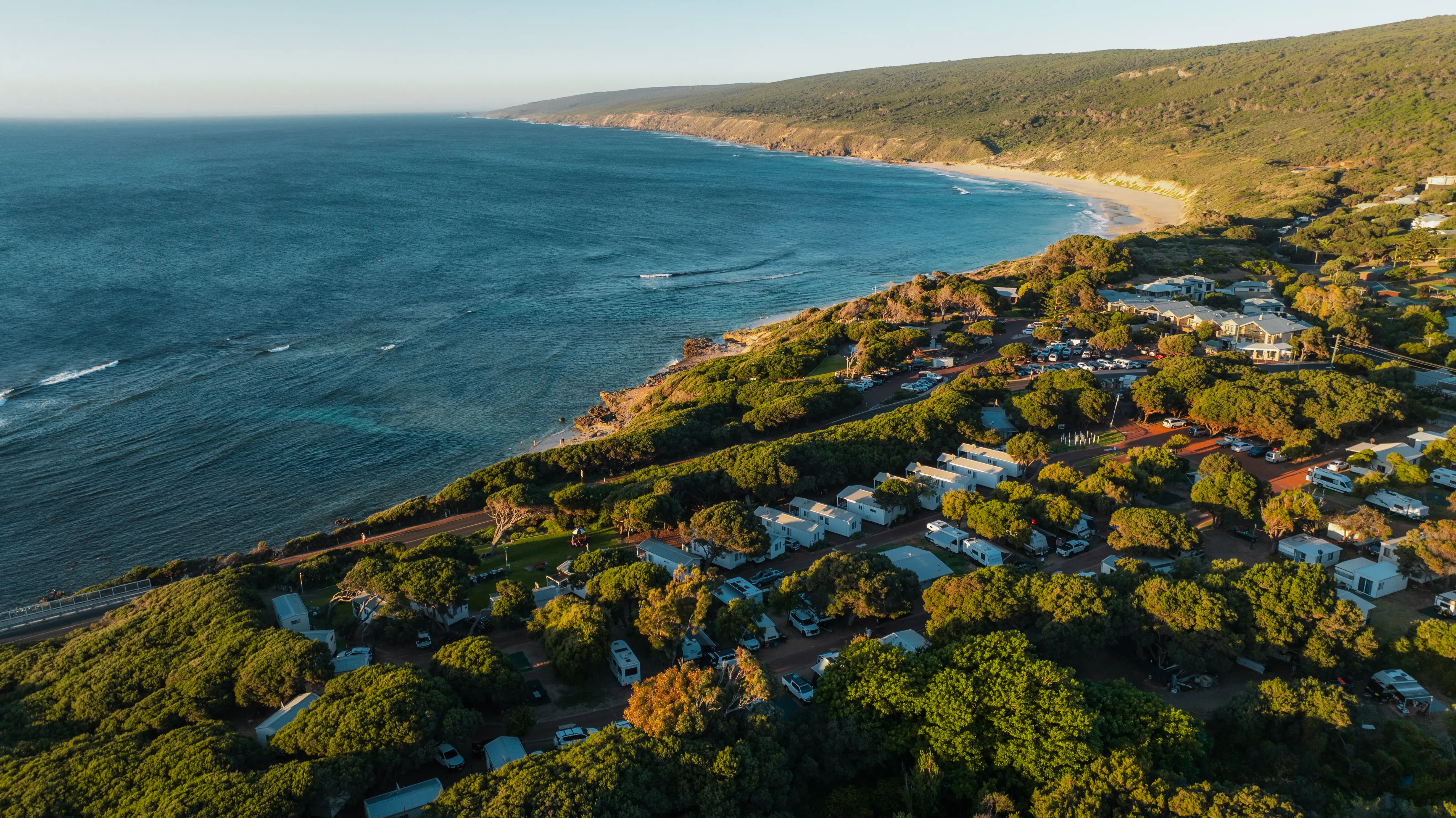 A coastal town with a blue ocean to the left, sandy beach and rocky shoreline, and a forested hillside dotted with houses, trees, and parked vehicles.