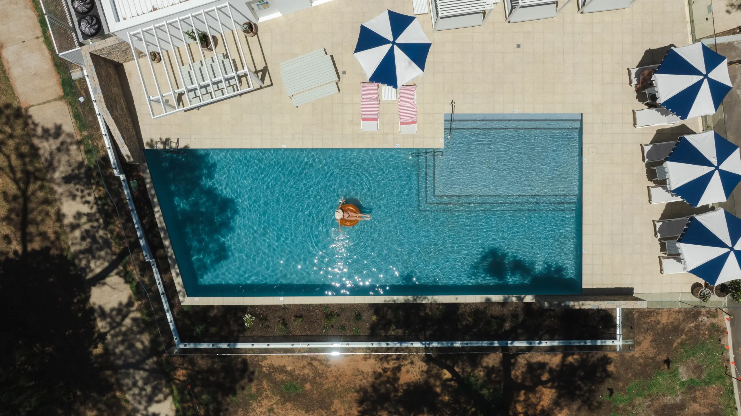 Aerial view of a rectangular pool area with turquoise water, a swimmer in the center, beige tiled deck, striped pink lounge chairs, blue and white umbrellas, and surrounding landscaping.