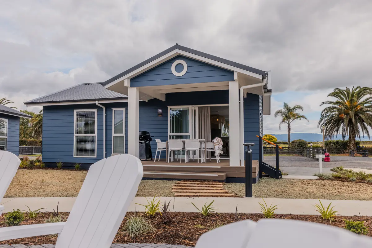Blue coastal bungalow with a covered front porch and white patio furniture; palm trees, gravel landscaping, and a wooden walkway lead to the entrance under a cloudy sky.