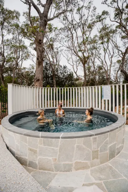 Three people soak in a circular stone hot tub outdoors, surrounded by tall leafless trees and a white railing, after a cool day.