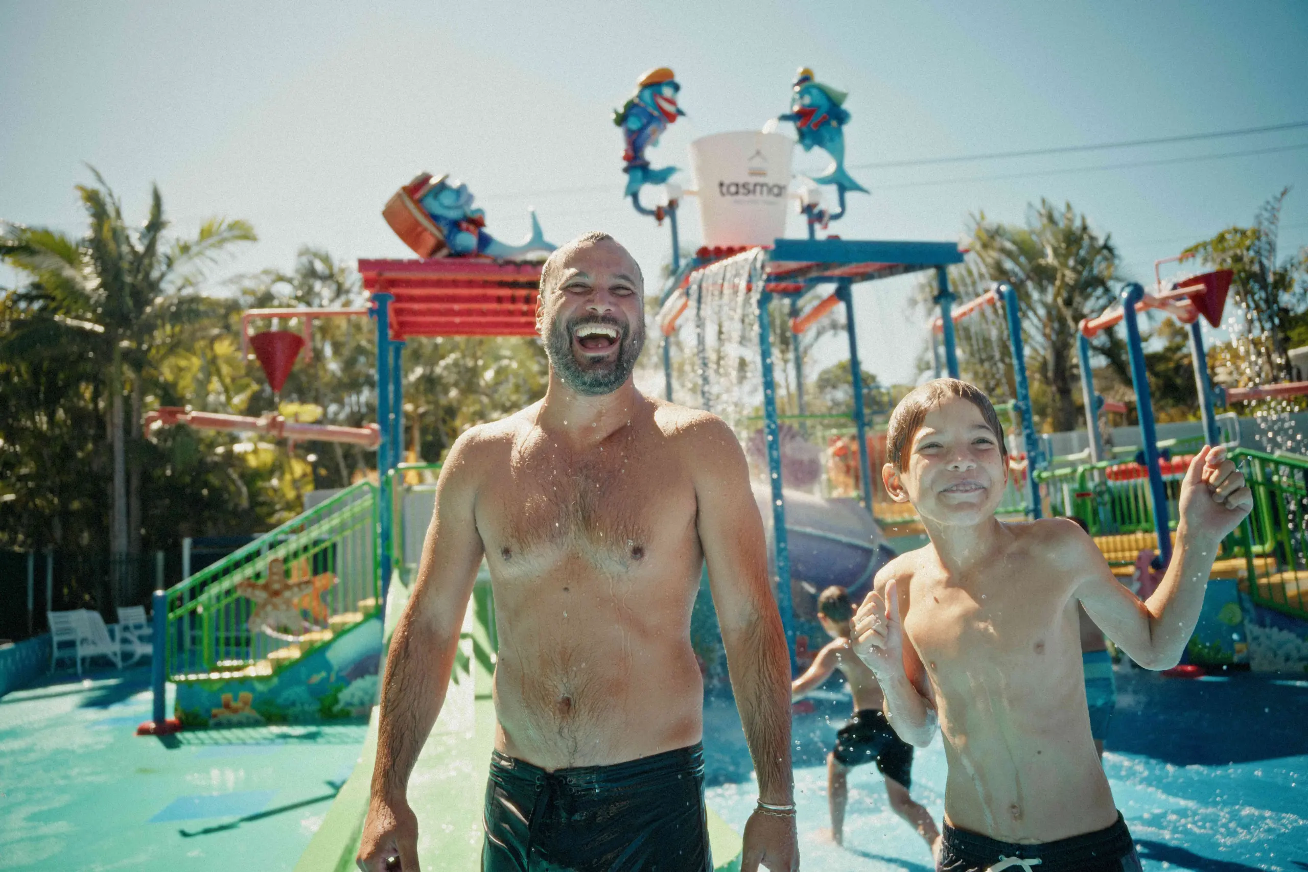 Two people, a bearded man and a boy, laugh and stand shirtless in a sunny water park with colourful slides and spraying fountains behind them.