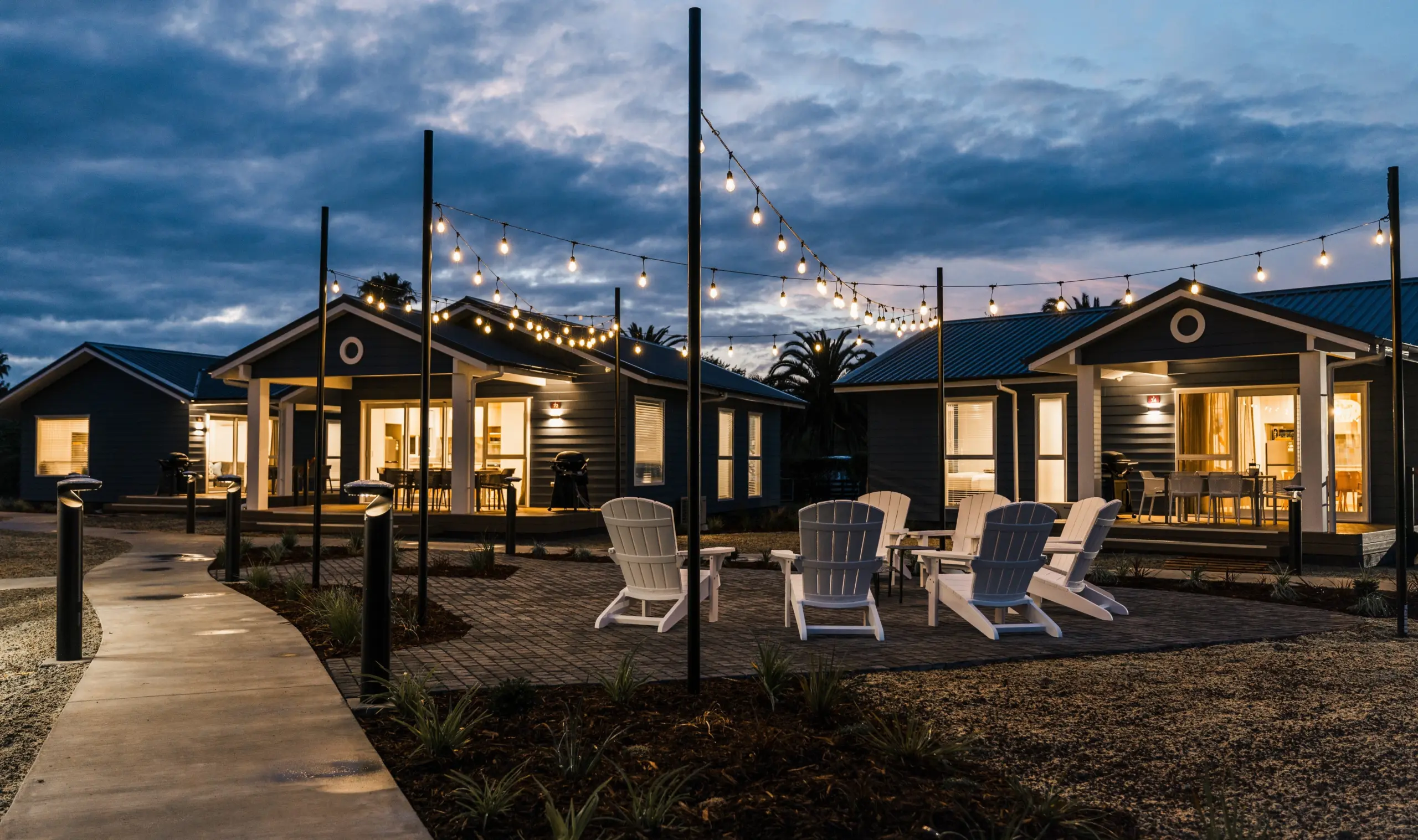 Outdoor modern cottages at dusk with string lights, a covered patio, and white Adirondack chairs surrounding a brick patio; warm interior lights glow from windows.