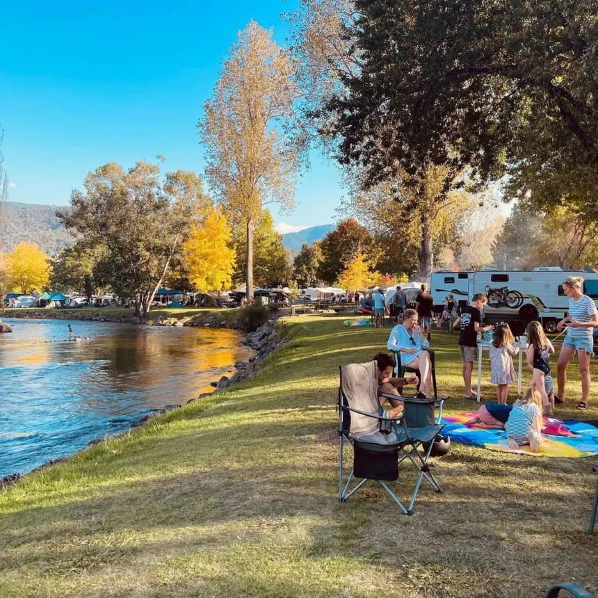 A sunny riverside park with people picnicking on blankets, trees in autumn colours, a lake on the left, tents and a camper van in the background, and mountains in the distance.