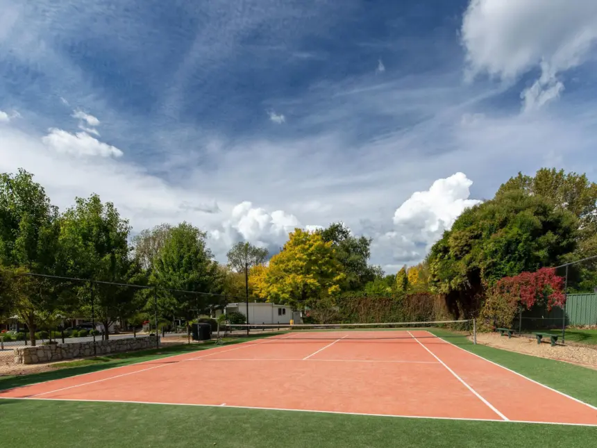 A clay tennis court surrounded by green grass, trees with autumn foliage, a clear blue sky with wispy clouds, and a fence-lined park area in the background.