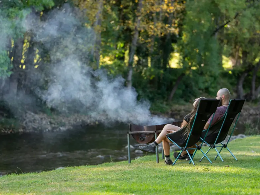 Two people relax on folding chairs by a river in a green park, smoke drifting from a nearby barbecue grill, with trees and dappled sunlight in the background.
