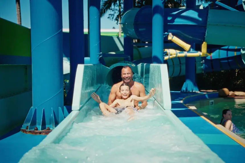 Two people slide down a blue water slide together, smiling as water splashes around, with colorful ride structures in the background at a sunny water park.