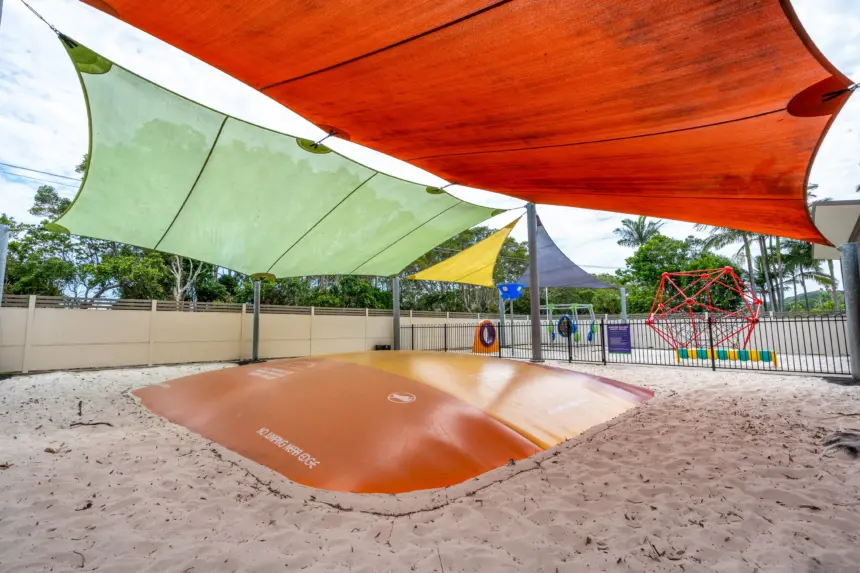 Outdoor playground area with large colorful shade sails (orange, green, yellow) over a sandy ground and a circular orange padded mound; fence and trees in background.