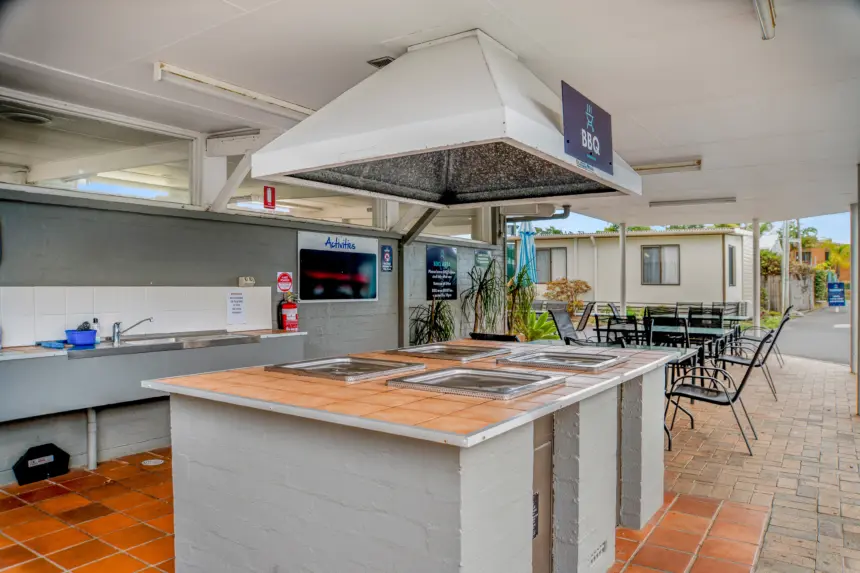 Outdoor BBQ area with a central stainless‑steel grill island, range hood, seating, and a cafe‑style patio under a covered walkway.