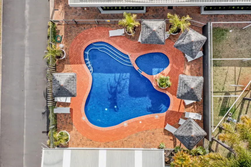 Aerial view of a curvy blue swimming pool with red brick around, sun loungers, thatched umbrellas, palm trees, and surrounding outdoor tiles.
