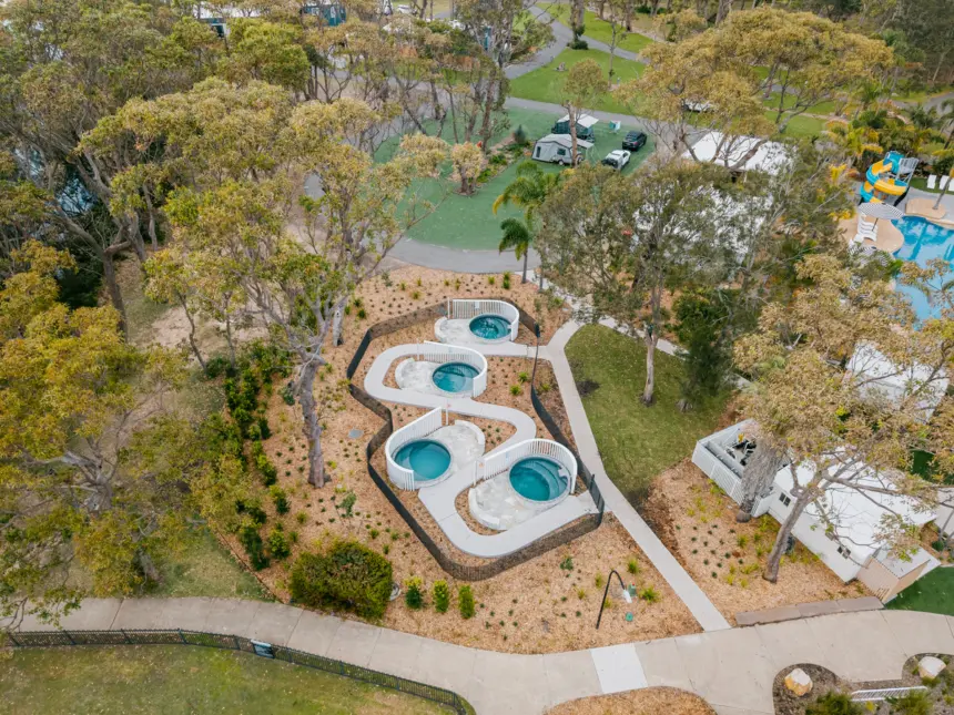 Aerial view of a landscaped communal area with four circular turquoise pools connected by curving white bridges, surrounded by trees, pathways, and amenities.