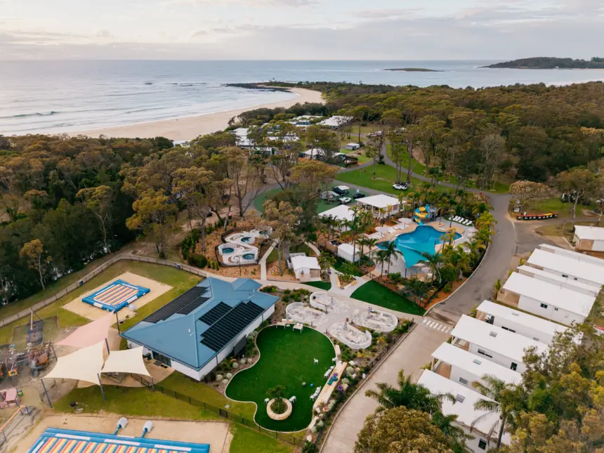 Aerial view of a coastal resort with a sandy beach, blue pool, green lawns, white buildings, shaded trees, and playgrounds, all bordered by a dense forest.
