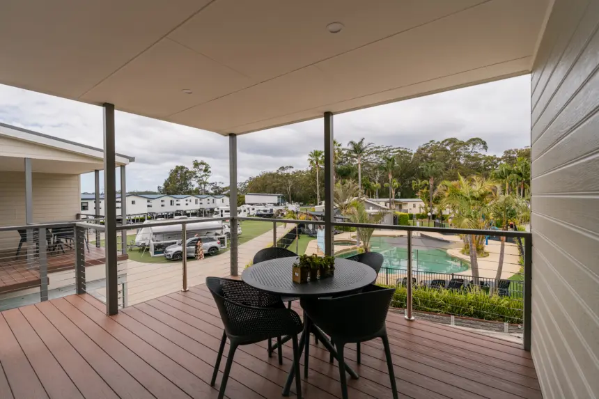 Balcony with round table and four black chairs overlooking a pool, palm trees, and a caravan park with mobile homes in a sunny, tropical setting.