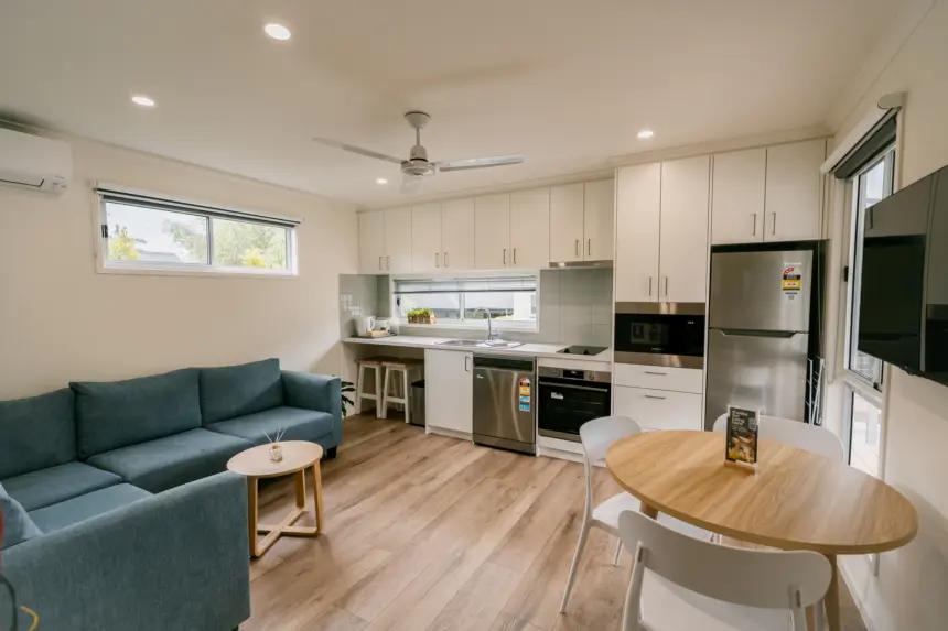 Open-plan modern kitchen and living area with white cabinetry, stainless appliances, light wood floors, a blue sofa, round dining table, and a wall-mounted TV.